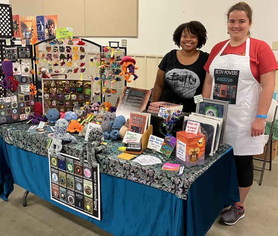 Two women standing behind a decorated table, featuring a book (Sorcery & Widgets), art items (mousepads, prints, stickers, magnets, keychains, pins), a drawing box, a poster, and a display of small and large crochet animals. 